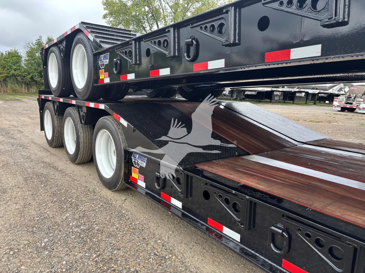 Lowboy trailer showing stacked flatbed sections with black painted frame, multiple axles, and white wheels; reflective tape along sides.
