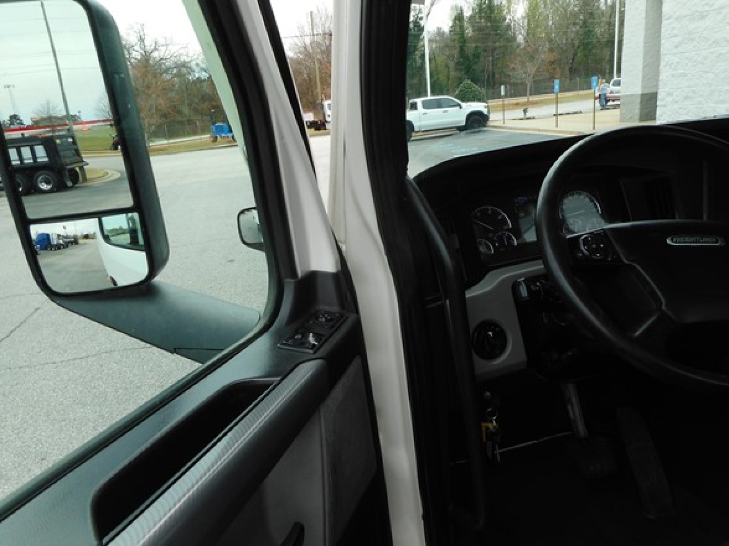 Truck cab interior with steering wheel, dashboard, and driver’s seat; exterior side mirror visible through open door window.