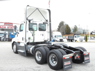 Truck rear view showing fifth wheel, dual rear axles, fuel tanks, and mud flaps. Visible text on mud flaps and license area; background includes other trucks and dealership lot.