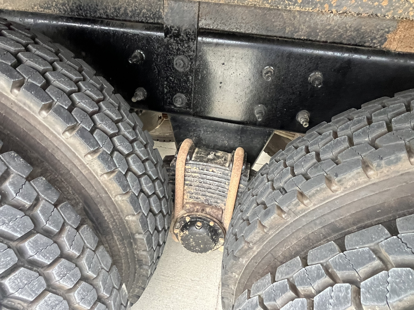 Truck axle area showing dual rear wheels and leaf spring shackle near frame rails, with visible bolts and grime.