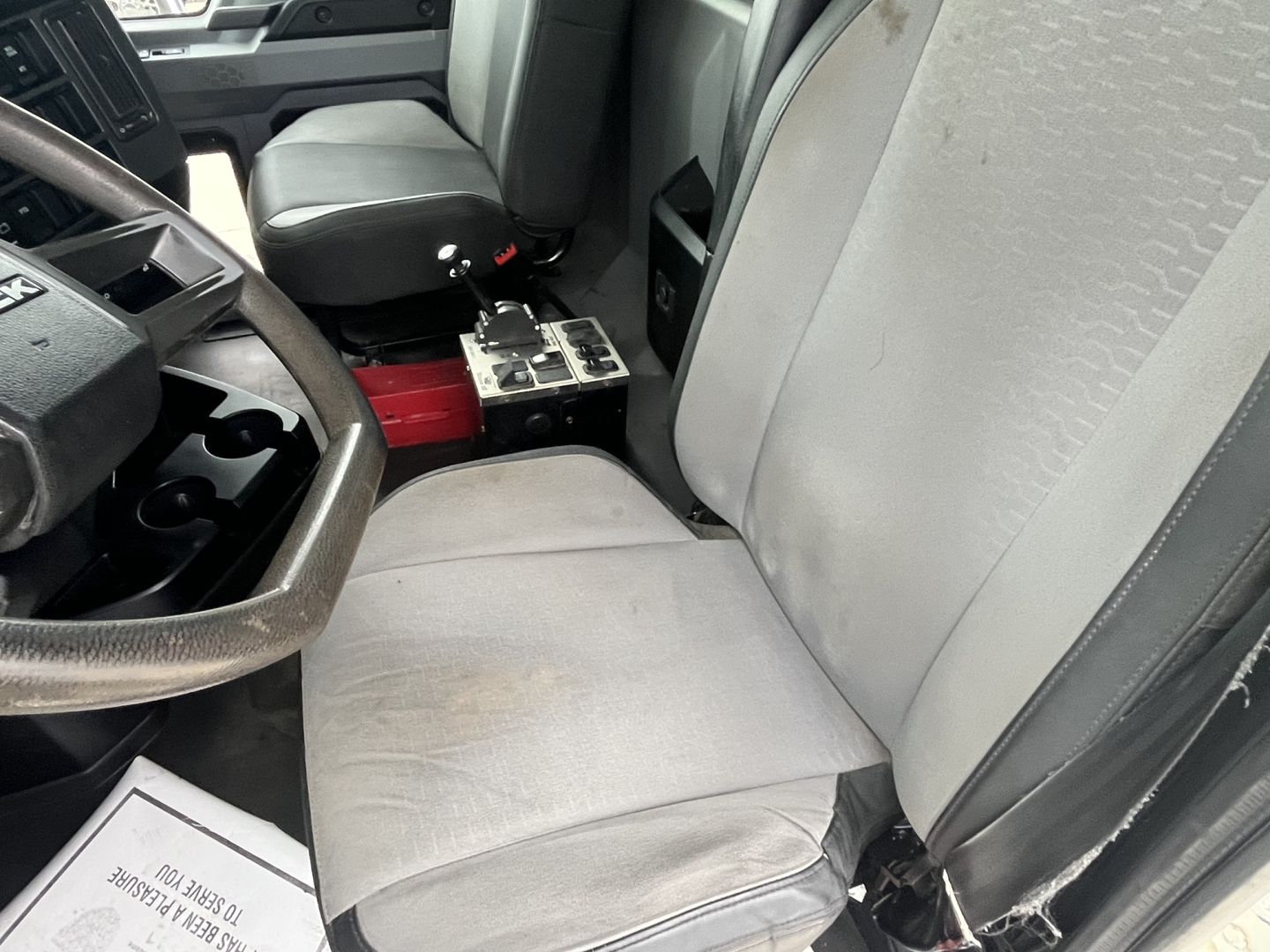 Truck cab interior showing driver seat and passenger seat with center console and exposed metal toolkit; steering wheel on left, cloth seats.