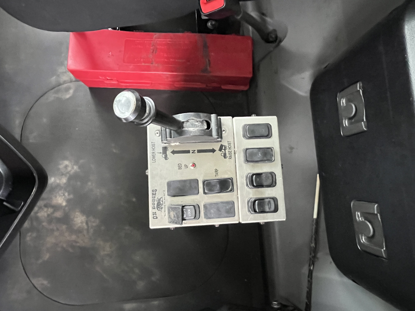 Cab interior of a 2020 Mack GR64F dump truck showing a joystick-style control module with several buttons and a lever, nearby red storage box and seat base.