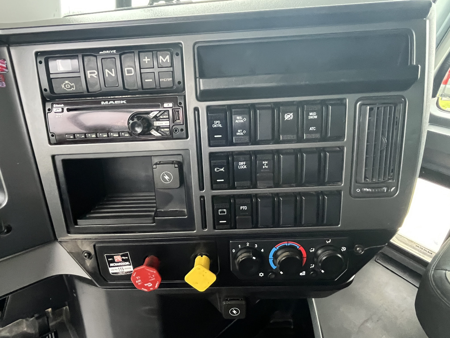 Mack truck dash showing gear selector buttons, radio, auxiliary switches, and climate controls; two colored control knobs (red and yellow) near lower center.
