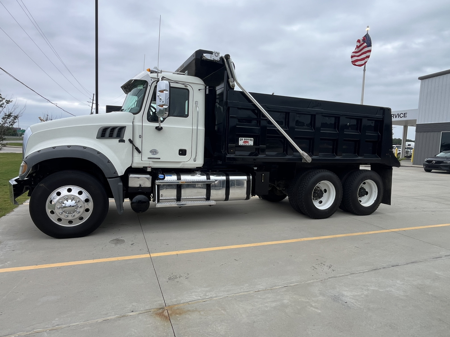 Dump truck side view showing 2020 Mack GR64F with white cab and black dump body, chrome fuel tanks, dual rear axles, and a raised tipping mechanism.
