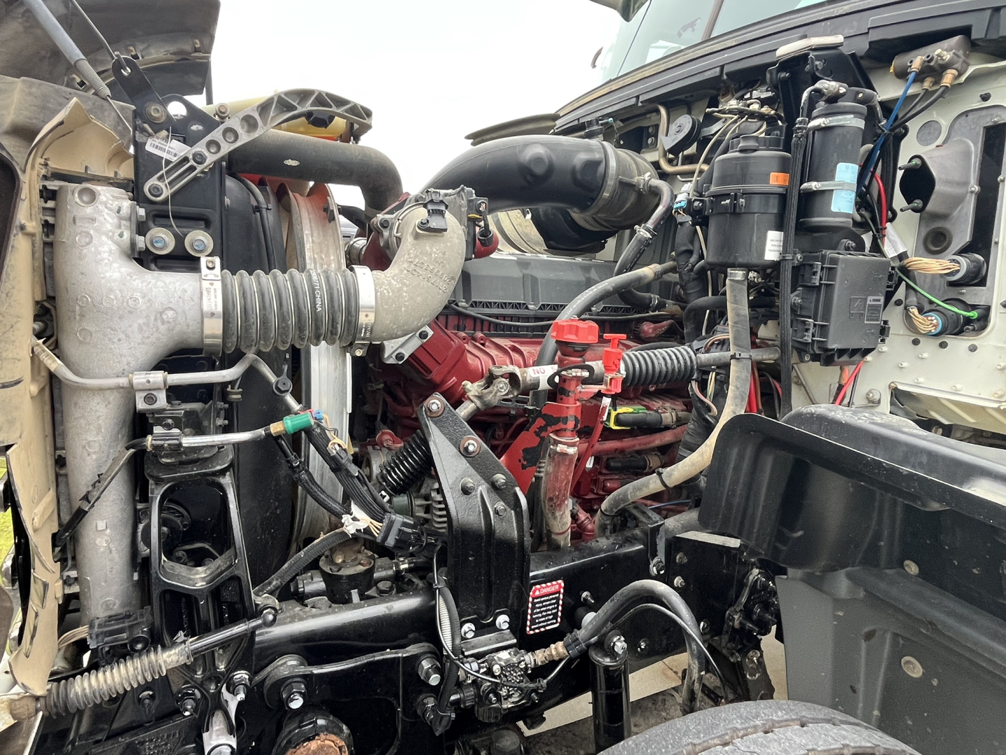 Truck engine bay with Mack GR64F diesel engine, hoses, belts, and electrical components visible; red engine block, air intake, cooling system radiator, wiring harnesses.