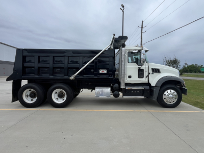 Dump truck side view showing 2020 Mack GR64F with white cab and black dump body, chrome exhaust stack, dual rear axles, and visible VIN plate area.
