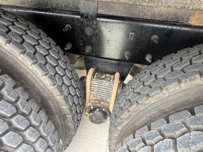 Truck axle area showing dual rear wheels and leaf spring shackle near frame rails, with visible bolts and grime.