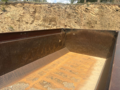 Dump truck bed interior showing rusted steel walls and orange-brown bed surface with dirt and wear.