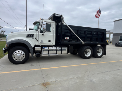 Dump truck side view showing 2020 Mack GR64F with white cab and black dump body, chrome fuel tanks, dual rear axles, and a raised tipping mechanism.