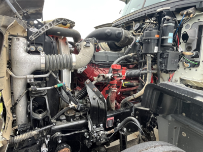 Truck engine bay with Mack GR64F diesel engine, hoses, belts, and electrical components visible; red engine block, air intake, cooling system radiator, wiring harnesses.