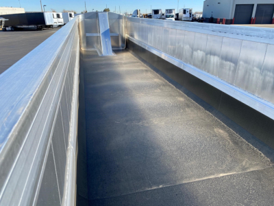 Dump trailer bed with metal side rails and flatbed surface, wide open cargo area, reflective aluminum panels, visible trailer yard in background.