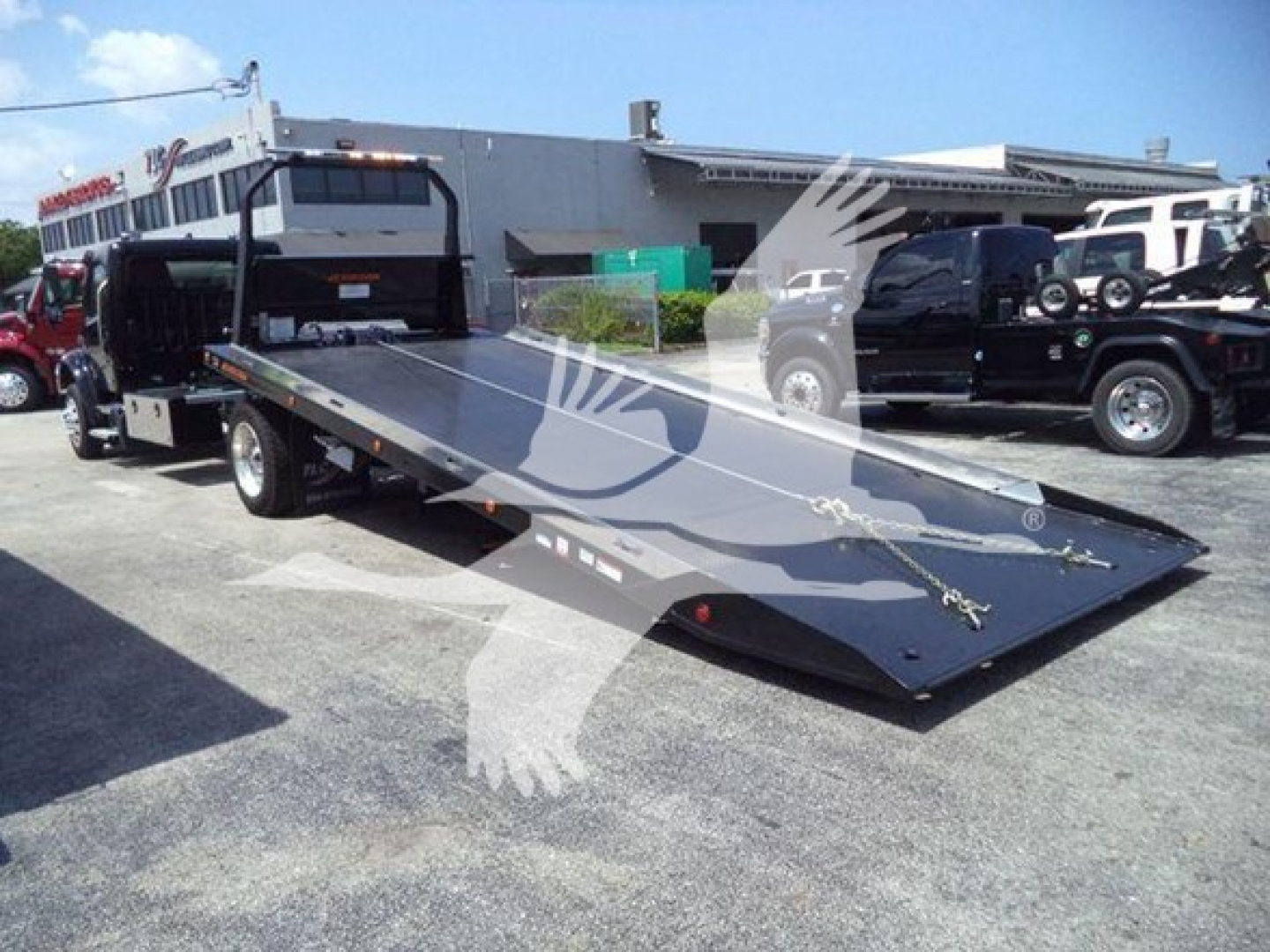 Flatbed tow truck with a lowered steel ramp bed extending from the back, parked in a lot with other trucks nearby.