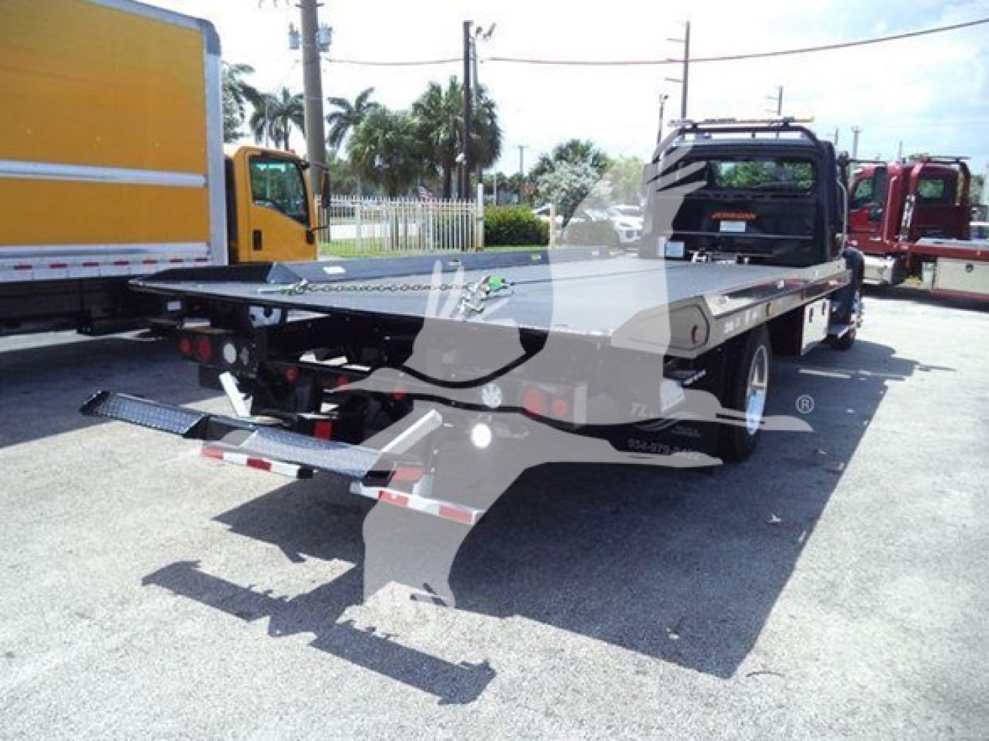 Flatbed tow truck with a hydraulic rollback bed, rear view showing chrome ramps and warning lights, Freightliner M2 106 Plus chassis in a dealership lot.