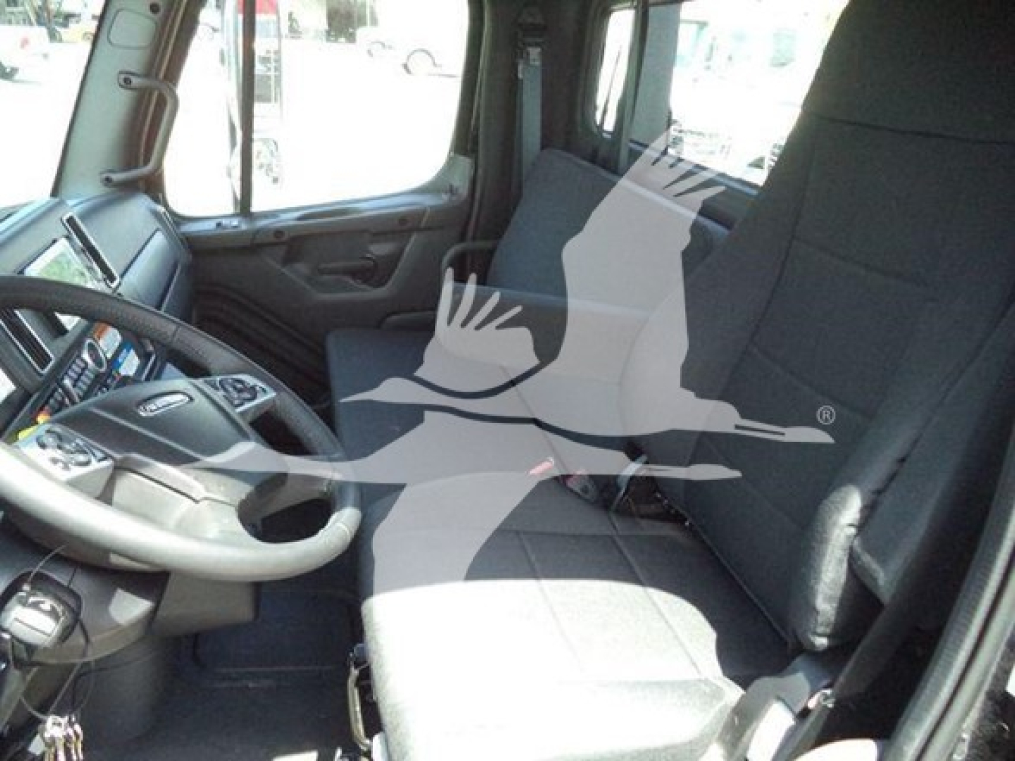 Freightliner M2 106 Plus truck interior driver seat and dashboard visible; steering wheel and control panel on left; cab seating area with large windows.