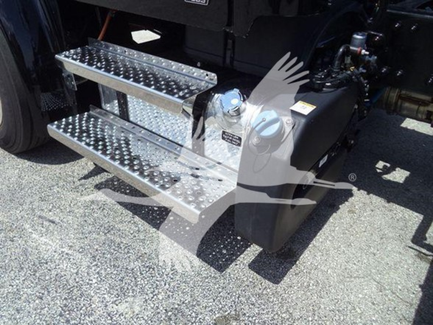 Step/run-flat steel steps with diamond tread beside Freightliner M2 106 Plus cab; fuel filler cap visible; hydraulic reservoir area; part of chassis and drivetrain.
