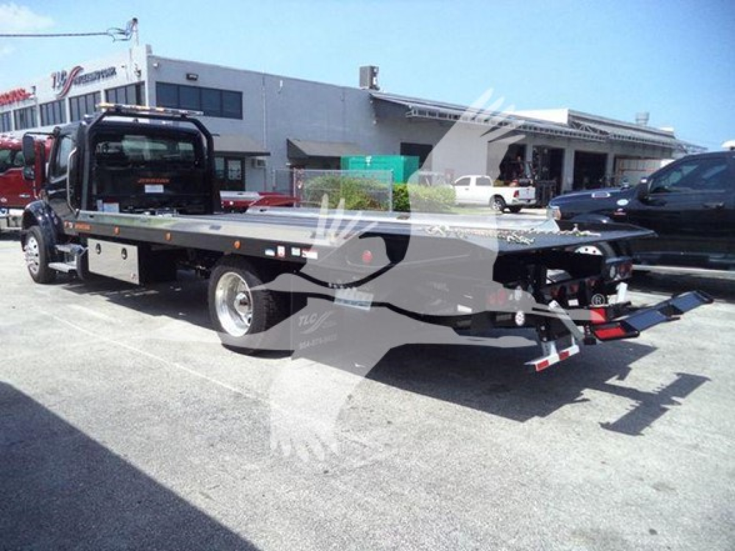 Flatbed rollback tow truck with extended bed and hydraulic ramps in a dealership lot; text on image includes no clearly legible detail due to watermark overlay