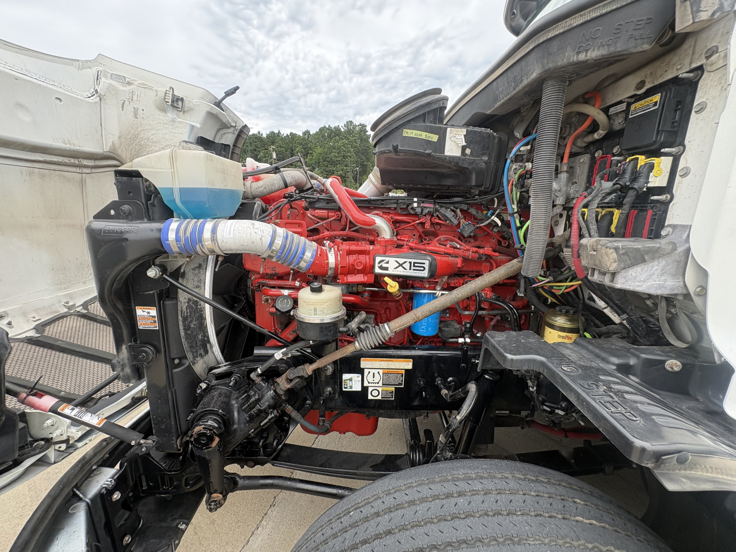 Peterbilt 579 engine bay showing Cummins X15 engine with red color, surrounding hoses, belts, and label stickers.
