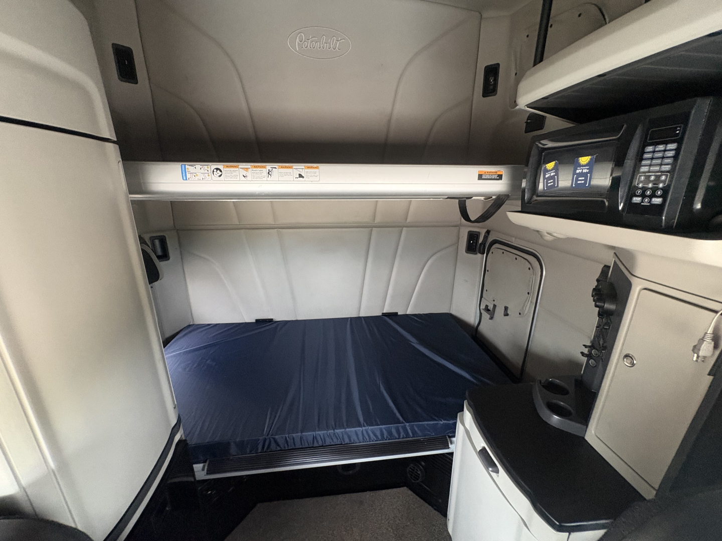 Peterbilt sleeper cab interior showing upper bunk area with mattress in blue cover, dashboard controls on right, shelves, and storage compartments.