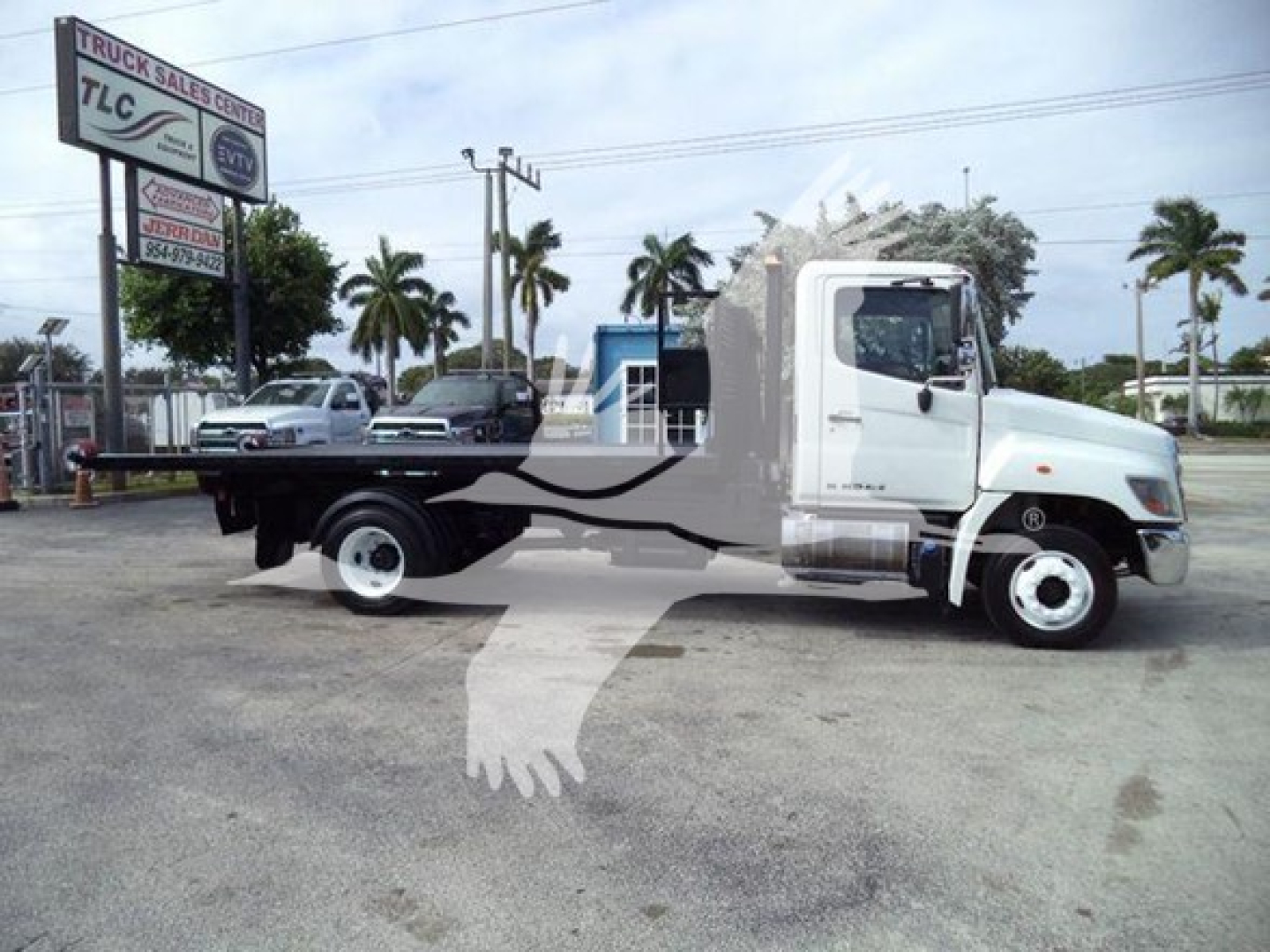 2017 Hino 258LP hooklift truck in a dealership lot with palm trees in background. Side view shows flatbed chassis and cab; TLC Truck Sales sign in distance.