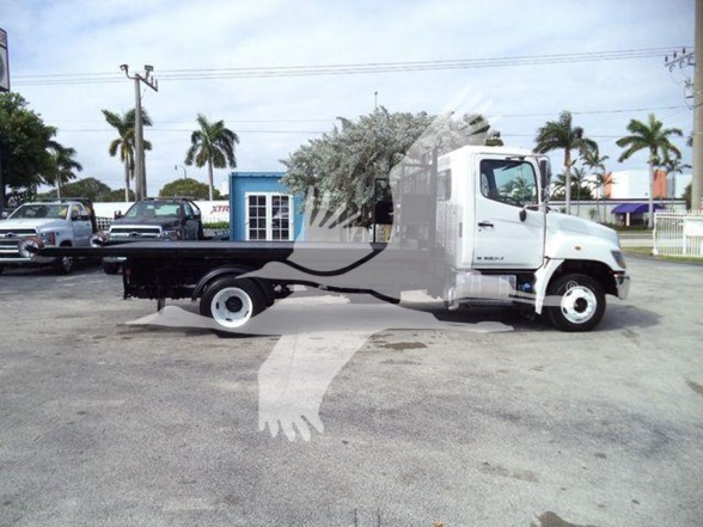 2017 Hino 258LP hooklift truck in a lot, cab white, long flatbed with black chassis, palm trees in background.