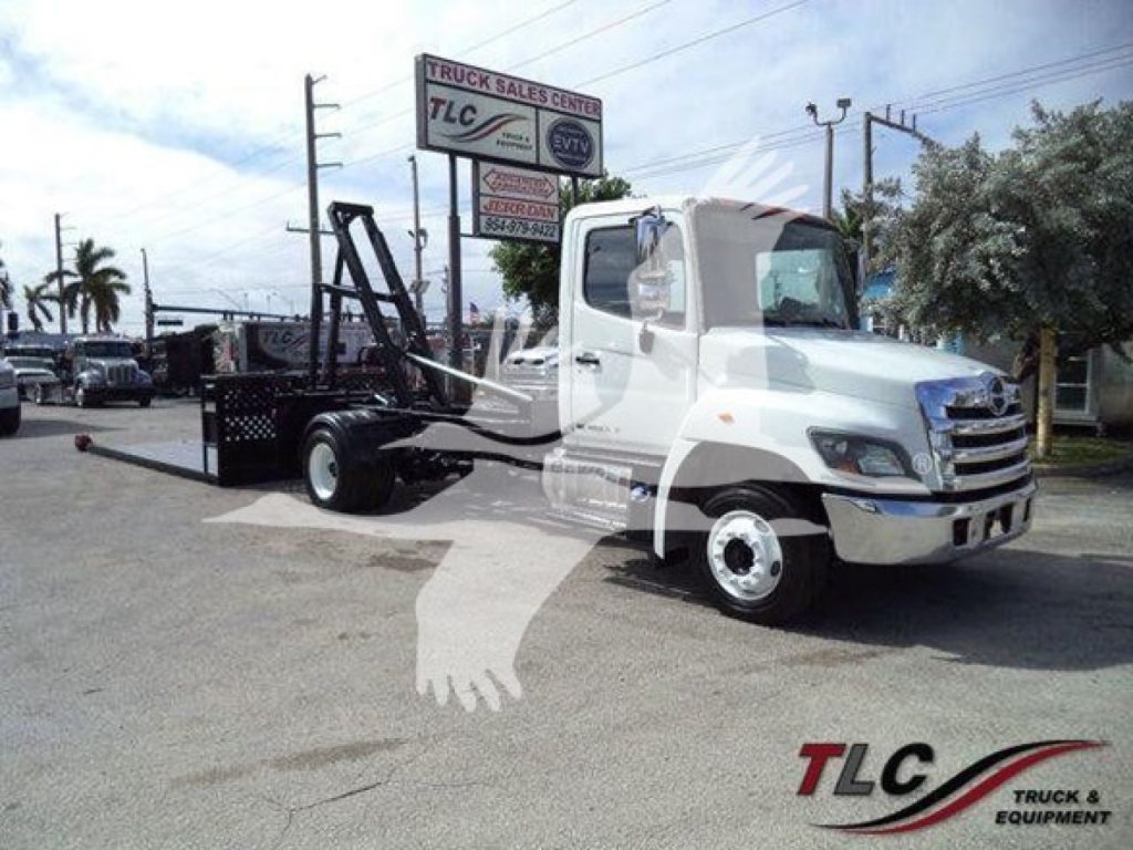Hino 258LP hooklift truck with equipment on the flatbed, dealership lot in background.