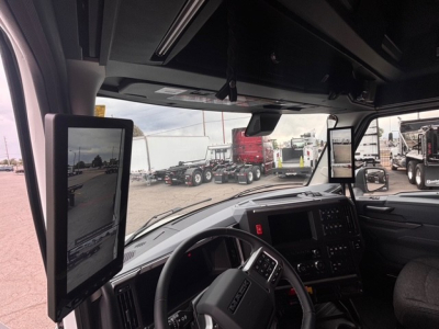 Cab interior of a Mack truck showing two mounted tablets on the A pillar and a steering wheel with dashboard; windshield view with trailers outside.