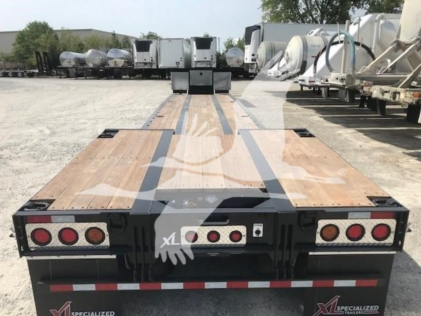 Lowboy trailer deck with wooden planks and red marker lights; rear view shows two sets of three red lights on each side and reflective tape, with 'XL' branding on tail; equipment yard in background.