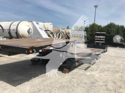Lowboy trailer showing flatbed deck with hydraulic ramps and dual-axle configuration; visible signage includes fuel/def tanks and chained tie-downs.