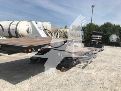 Flatbed trailer in a salvage lot showing a long lowboy deck with ramps, dual rear axles, and a truck bed; some tanks and equipment in background.