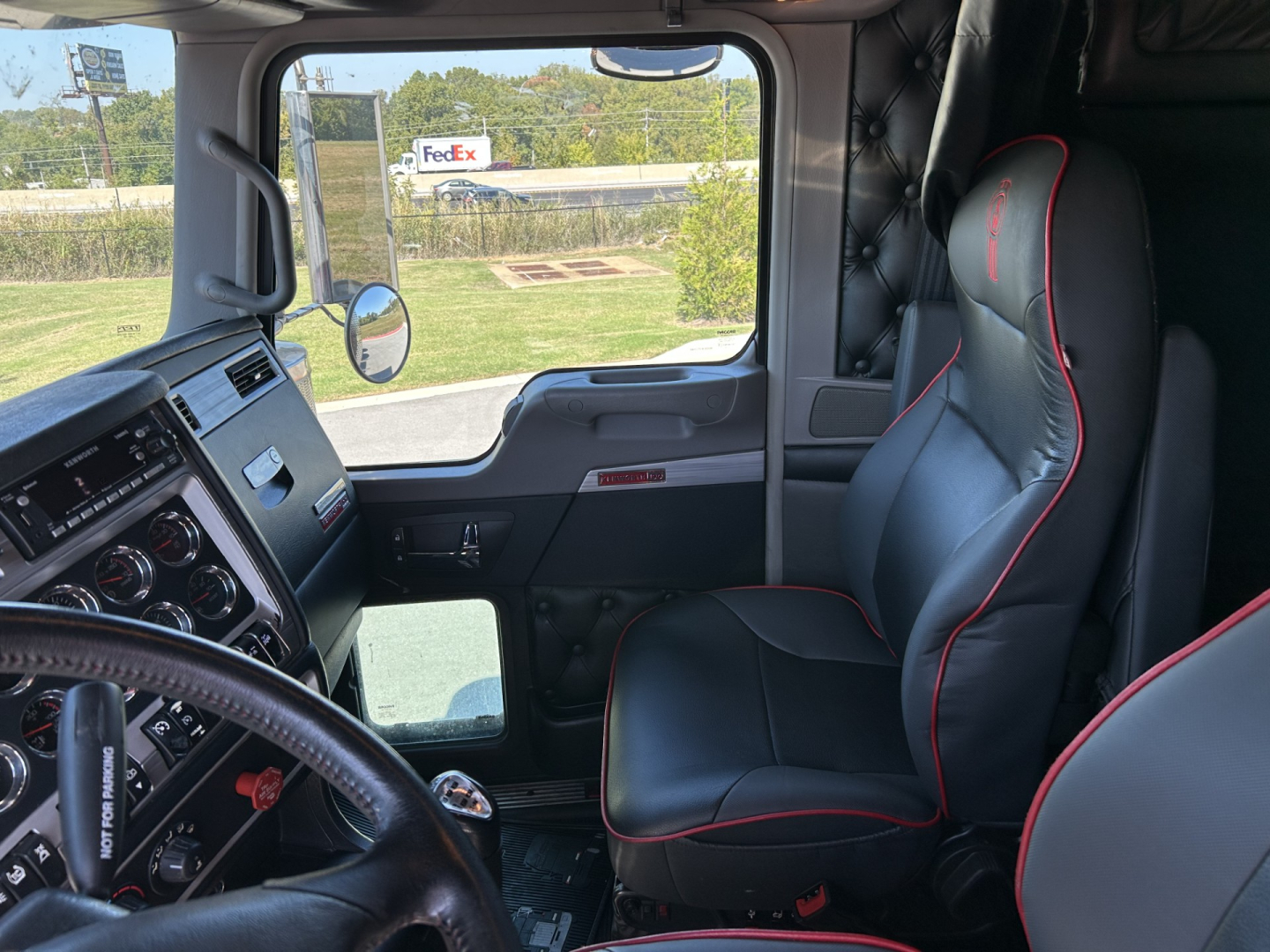 Interior of a Kenworth W900L truck cab showing steering wheel, dashboard, and black leather seats with red piping.