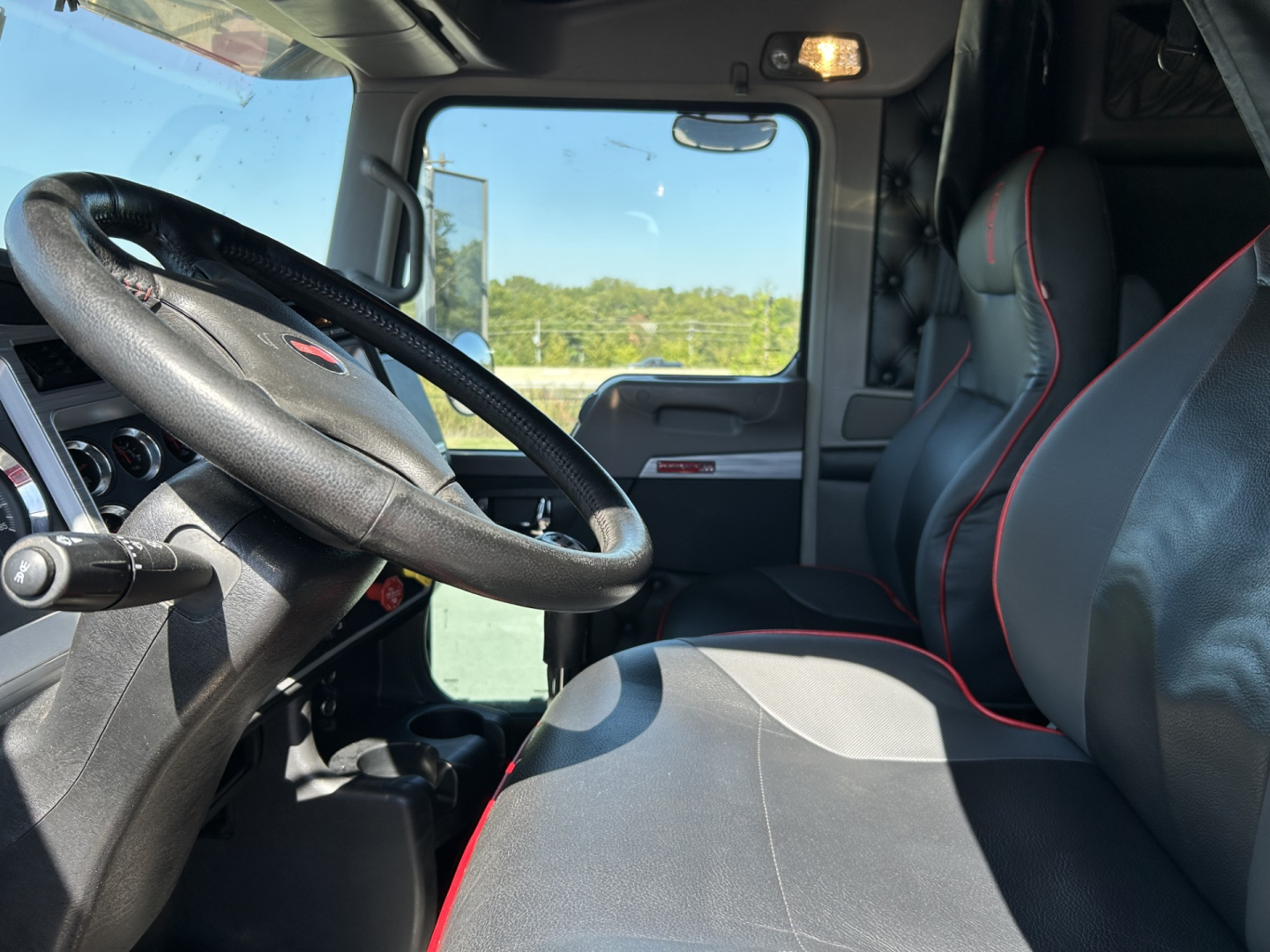 Kenworth W900L interior cab view showing steering wheel, driver's seat with black and red trim, dashboard, and door panel.