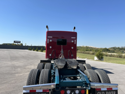 Kenworth W900L chassis view showing rear section and fifth wheel, blue frame with dual rear axles and red sleeper cab in background.