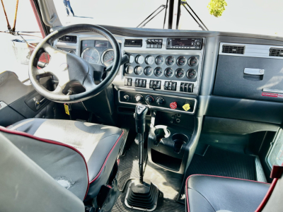 Dashboard and steering wheel of a 2024 Kenworth W900L showing instrument cluster, gear shifter, center console with numerous gauges and controls, and steering wheel with Kenworth logo.