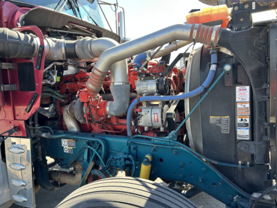 Kenworth W900L engine bay showing red diesel engine, blue chassis, large gray coolant hose, radiator, and various hoses and belts.