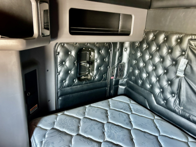 Interior sleeper area of a Kenworth W900L cab showing diamond-stitched upholstery, storage cabinets, and window opening with a mounted control panel.