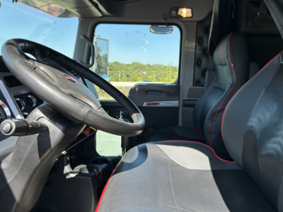 Interior of a Kenworth W900L truck cab showing steering wheel, driver seat with black and red trim, and passenger seat.