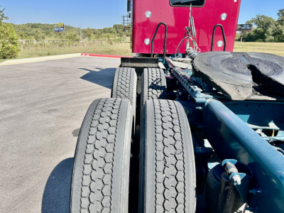 Kenworth W900L truck chassis view showing tandem rear axles with dual tires and part of red sleeper cab in background.