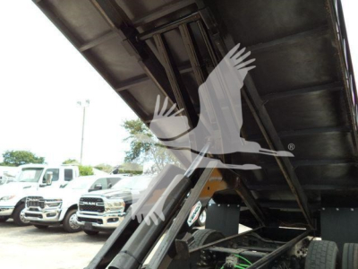 View under a dump truck bed showing hydraulic linkage and wheel assembly; visible stickers include 'GLOBE' or similar logo on a black hydraulic component. Various trucks parked in background.