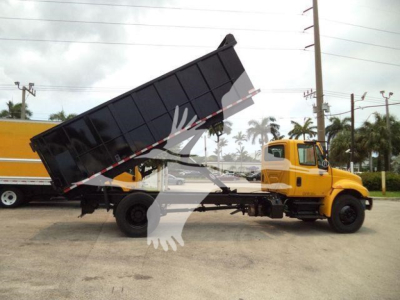 Truck side view: yellow International 4300 dump truck with raised black dump bed. Weathered pavement and palm trees in background.