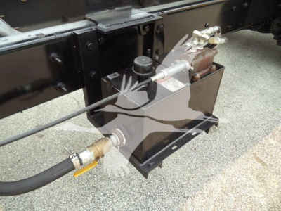 Truck chassis with hydraulic reservoir and hose assembly mounted near frame rail; black metal box with fittings and a hydraulic line fitting.