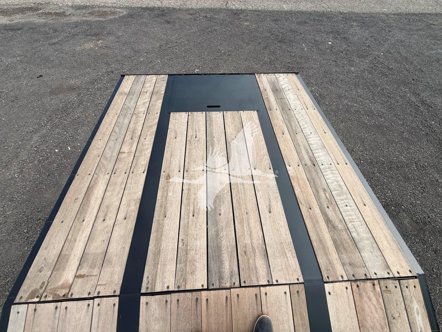 Trailer bed showing wooden deck planks with a central dark metal panel and a small access hatch, viewed from above.