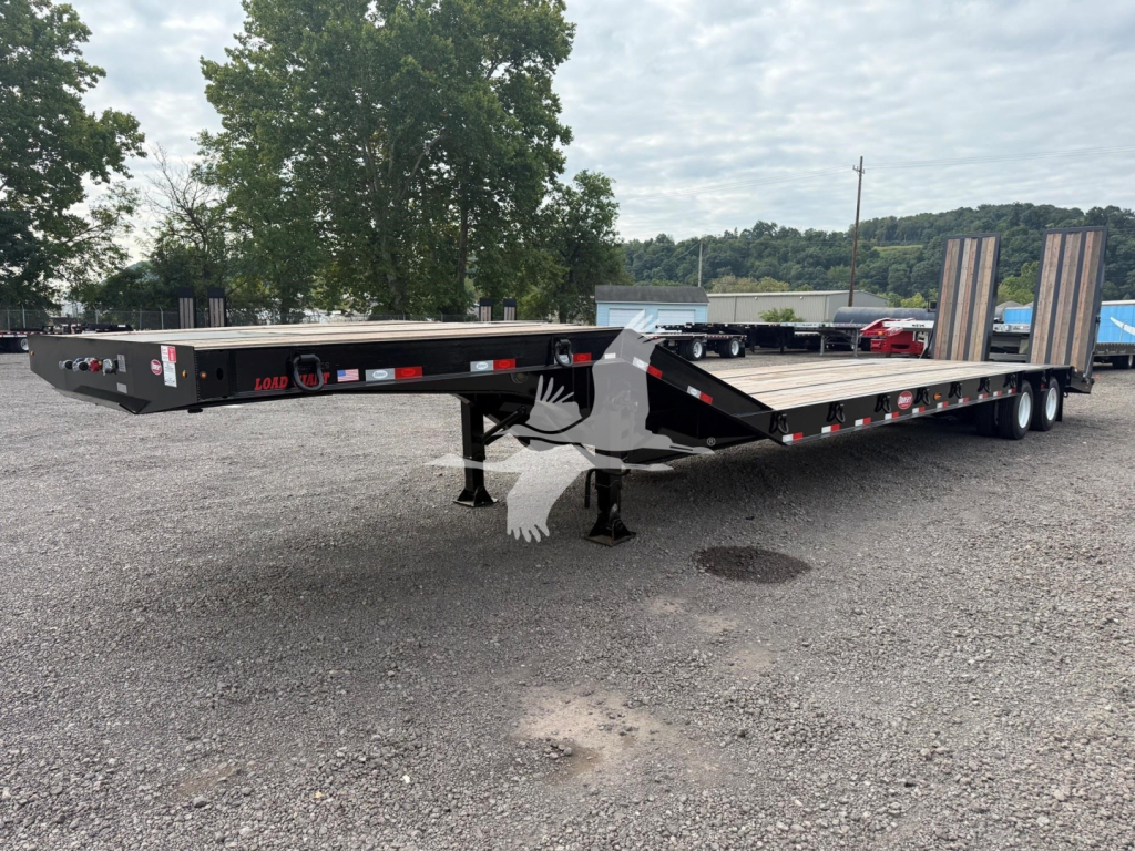 Trailer in a parking lot showing a long flatbed drop deck trailer with dual rear axles and ramps, brand labels on side, and support legs deployed.