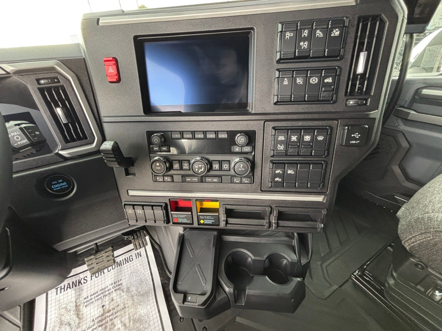 Dashboard of a 2026 Mack PR64T76H showing control panel, infotainment screen, climate and switch panels, cup holders, and storage compartments.