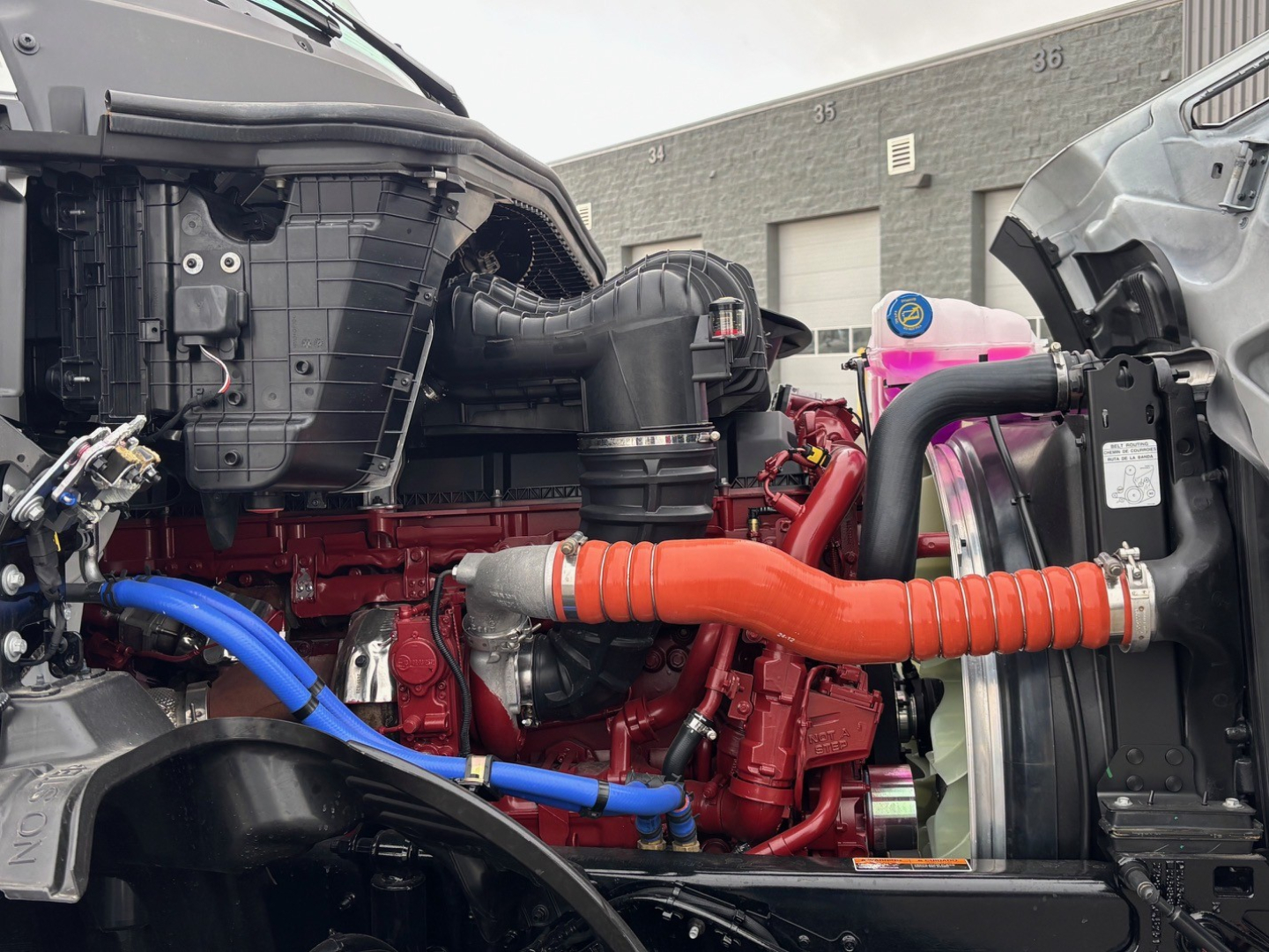 Mack PR64T76H drivetrain with red engine block, orange coolant hose, blue hoses, and exposed components in engine bay.