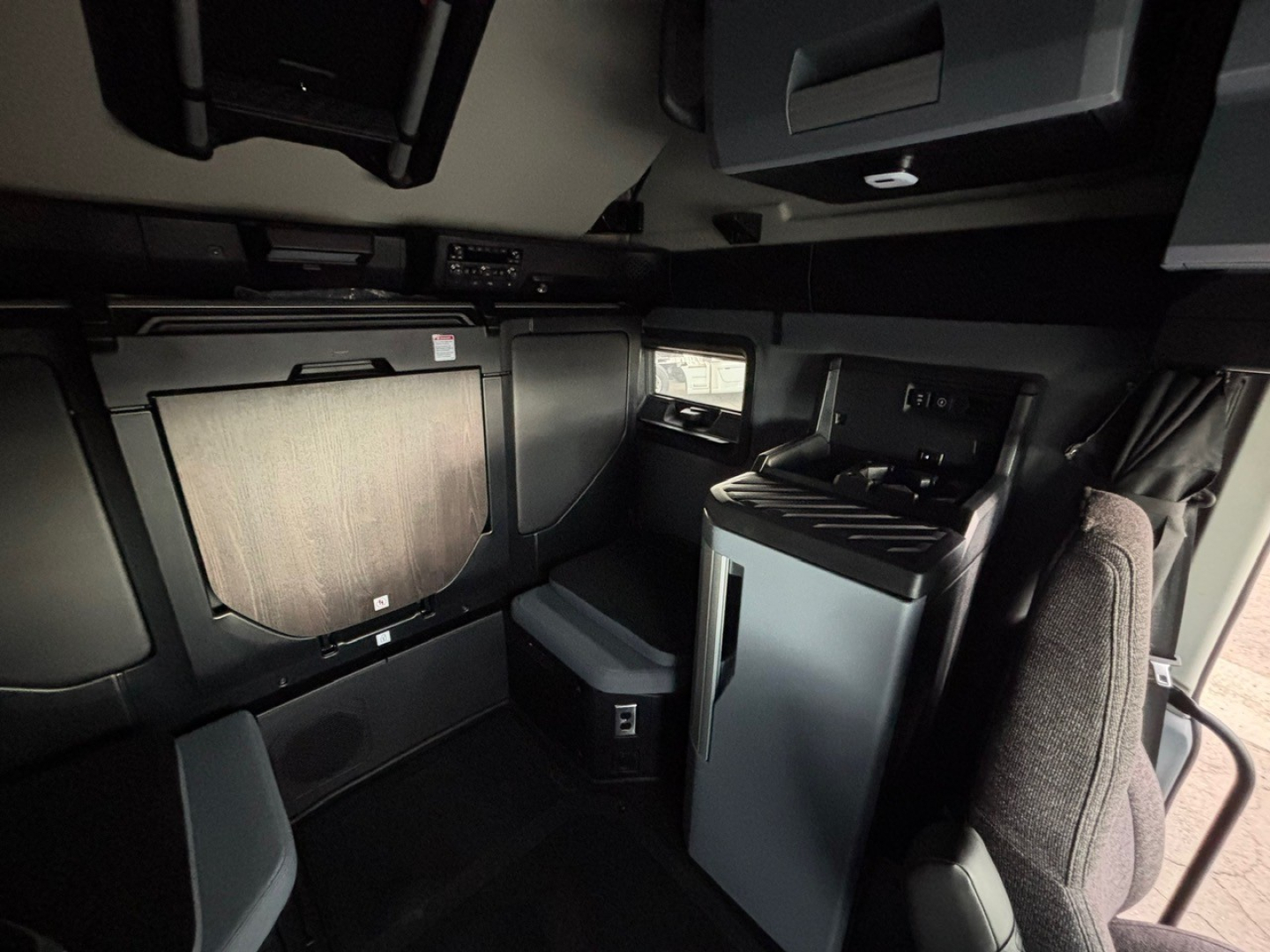 Interior cab view of a new Mack PR64T76H sleeper truck showing a compact living area with cabinets, a small fridge, and driver seat in the foreground.