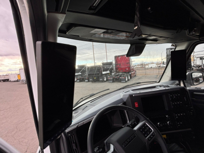 Truck cab interior with dashboard and steering wheel, view through windshield showing other trucks in lot.