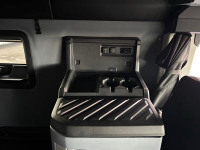 Cab interior showing storage compartment and control panel in a Mack PR64T76H sleeper tractor, with a center console and power outlets visible.