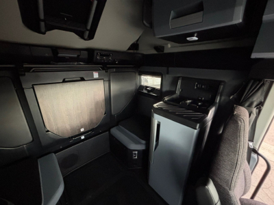 Interior cab view of a new Mack PR64T76H sleeper truck showing a compact living area with cabinets, a small fridge, and driver seat in the foreground.