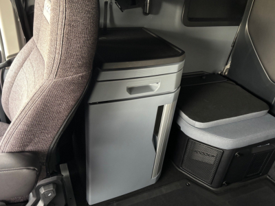 Cab interior of a new Mack PR64T76H truck showing a grey under-dash cabinet with a drawer and a vertical storage compartment, adjacent to a seat and a secondary storage box.