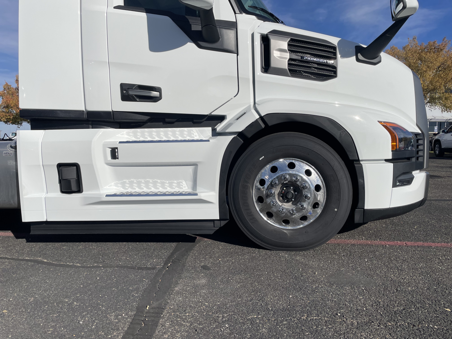 Mack PR64T truck left-front section with wheel, fender, and steps visible; white cab and chrome wheel hub; blue sky in background.